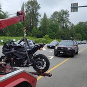 A black motorcycle is towed to the impound yard as a BC Highway Patrol cruiser is visible in the background