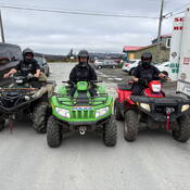 Two uniformed RCMP officers and one uniformed Conservation Enforcement Officer are geared up and heading out on ATV patrol. Marked police vehicles and a search and rescue command post vehicle are seen in the background.