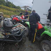 Two uniformed RCMP officers are preparing their gear to head out on ATV patrol, one is pouring gas into a green ATV from a jerry can.