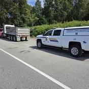 A large dump truck hooked up to a red tow truck on the side of the road, with a police commercial vehicle inspection pick up truck parked behind it.