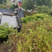A view from the top of the embankment. The SUV can be seen on top of the white wooden shed. Bushes and trees surround the heavily damaged shed.