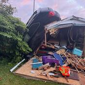 The SUV’s rear tires are on the roof of the shed, and its front bumper rests on the ground. The whole rear wall of the shed is collapsed and the damaged contents of the shed are strewn all over.