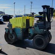 A yellow and green Ammann steamroller, viewed from the side, is stopped next to an orange traffic pylon in a parking lot. No one is on the steamroller and vehicles are visible in the background.