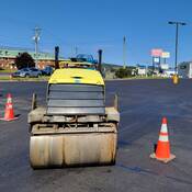 A yellow steamroller, viewed from the front, is stopped between orange traffic pylons in a parking lot. No one is on the steamroller and businesses, and vehicles are visible in the background.