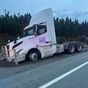 A transport truck is parked on the gravel shoulder of a roadway with its front end dug into the gravel and a flattened front driver side tire.