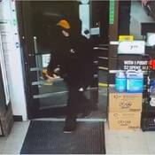 An individual steps inside the store. The person is wearing a black coat, a pair of black pants, a pair of black and white shoes, a black hoodie, a pair of black and red work gloves and a yellow/orange base ball cap;