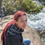Photo of Ashley Bosma, apparently Caucasian woman, 40-years old, crouched on a rock near rushing water and looking up at the camera. She has long bright red hair pulled back into a pony tail and is wearing a hoodie with a diamond pattern hood.