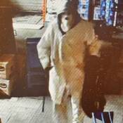 A still image of the suspect taken from video surveillance. The suspect is carrying a black duffle bag and is seen wearing a full-face mask, a white coat, white pants and has a hood up.