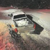 a light-colored GMC pickup truck is stuck in deep snow, off the side of a snow covered roadway. Trees are visible in the background.