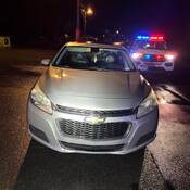 A grey Chevrolet car is seen on the side of the roadway with a police SUV behind it. The police vehicle’s red and blue emergency lights are on.