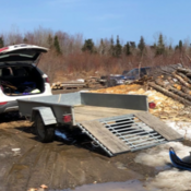 An aluminum utility trailer - with its ramp down - is attached to a white SUV. The hatch of the SUV is open and the trailer is empty;