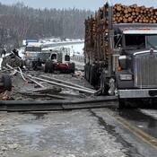A logging truck is stopped on a snow and ice covered bridge deck amongst crash debris. A heavily damaged white commercial tractor truck is visible in the background.