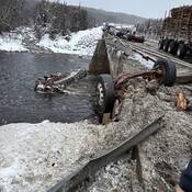A damaged tanker trailer is in the waters of a river below a severely damaged bridge deck. A damaged tractor truck is on the bridge deck amongst crash debris.