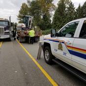 A police officer wearing a yellow vest ensures a load is safely strapped down in front of a police vehicle, while a truck driver stands beside his dump truck.