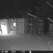 A person stands using a tool, attempting to break into a shipping container in the darkness. Sparks can be seen.