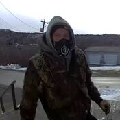 A surveillance image of a masked man – standing on a wooden deck – wearing a camouflage jacket, a grey hoodie and gloves.