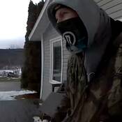 A surveillance image of a masked man - stands outside a home – wearing a camouflage jacket, and a grey hoodie.