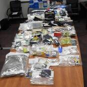 A large number of items are displayed in clear evidence bags on a table, including firearms, cash, pills, drugs, brass knuckles and contraband tobacco.