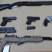 A shot gun, a semi-automatic rifle, three handguns and four magazines of ammunition are displayed on a table.