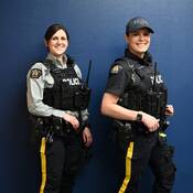 Two RCMP officers in uniform, one wearing the long-sleeved grey shirt, the other the short sleeved new navy-blue uniform shirt.