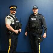 Two RCMP officers in uniform, one wearing the short sleeved grey shirt, the other the long-sleeved new navy-blue uniform shirt.