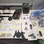 Various forms and quantities of illicit drugs, a sum of cash, contraband tobacco, multiple firearms, ammunition and a set of brass knuckles are displayed on a table. Pieces of paper, one shows the RCMP and RNC crests with “JFO West” in writing, another show the RCMP crest with “West District GIS” in writing, and another shows the RCMP Crest with “Newfoundland RCMP Rocky Harbour Detachment” in writing.