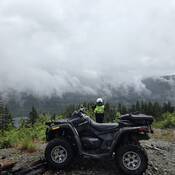 Photo of a police ATV with an RCMP officer standing nearby, set against the backdrop of mountains covered in cloud