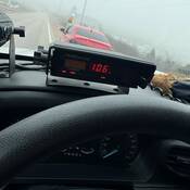 A red-coloured car stopped on the side of the road. In the foreground, a police radar shows a speed reading of 106 km/hr.