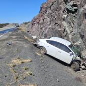 A heavily damaged white passenger vehicle is in the ditch and rests up against a rock wall. Transport trucks and other motor vehicles are visible in the background.