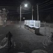 Two individuals, dressed all in dark clothing with hoods over their heads, walk towards an open gate dragging large coils of wire.