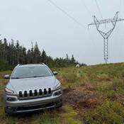 A grey Jeep SUV sits below transmission lines, with a marked police vehicle visible in the distance.