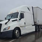 A white Freightliner commercial tractor is partially off the roadway, and its attached white trailer fully blocks one lane of the two-lane roadway.