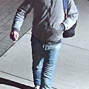 Photograph of male walking on sidewalk, wearing a hoodie, blue jeans, carrying a backpack.