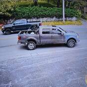 a four-door pickup truck captured on a surveillance camera. The truck appears grey and light blue, has a sunroof and items in the back, including a lawn mower.