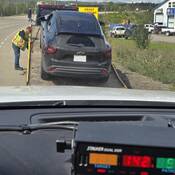 A tow truck driver loads a black Chevrolet Trax onto his truck while a BC Highway Patrol dash-mounted radar shows a reading of 142 km/h