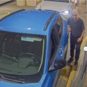 A man stands next to the driver side door of a blue vehicle facing the surveillance camera.