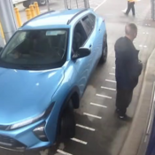 A man, dressed in dark clothing, stands next to the driver side of a blue vehicle and faces away from the camera.