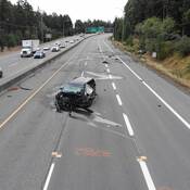 A Kia minivan with severe front-end damage in the southbound lanes of Island Highway North, with a heavily damaged Volkswagen Jetta in the ditch in the background