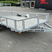 An aluminum/silver tandem axel utility trailer, with a wooden deck is parked in a lot. The words “EASY HAULER” are om black lettering on the side of the trailer’s tongue.