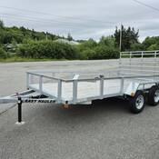 An aluminum/silver tandem axel utility trailer, with a wooden deck is parked in a lot. The words “EASY HAULER” are om black lettering on the side of the trailer’s tongue.