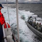 A uniformed member of the Canada Coast Guard stands on a vessel and looks on as a grey RCMP Zodiac vessel with four RCMP Emergency Response Team (ERT) officers on board pulls alongside.