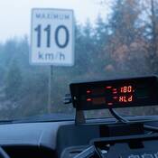 A radar reader shows 180 on the dashboard of a BC Highway Patrol cruiser. The cruiser sits in the rain in front of a speed limit sign that reads 110 km/h