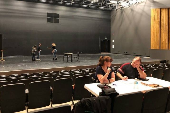 A man and a woman sit talking with their backs to actors on a stage. 