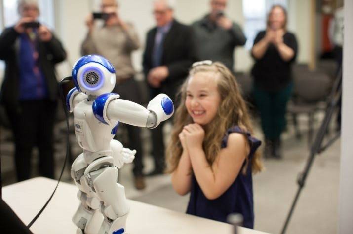 Young girl stands gleefully watching a two-foot-tall white and blue robot standing on a table.