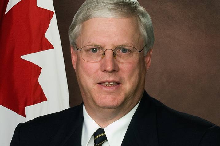 Portrait of a man wearing a dark suit and tie, with a Canadian flag behind him.