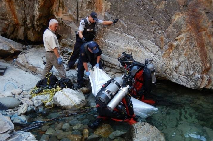 Two RCMP divers pull a bagged drowning victim from the water while male officers retrieve it from a rocky shore.