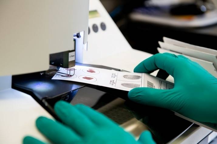 Gloved hands directing a card with blood samples and fingerprints impressions on it through a sampling machine in a lab.