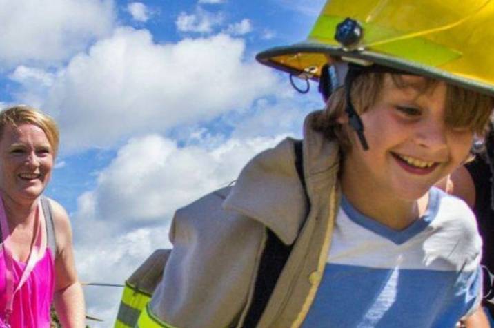 A young boy wearing a firefighter's jacket and helmet runs and smiles. A woman stands behind him smiling.