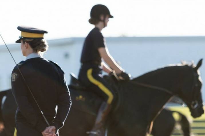 A female RCMP officer stands in a riding ring, holding a crop. She watches other officers ride their horses.