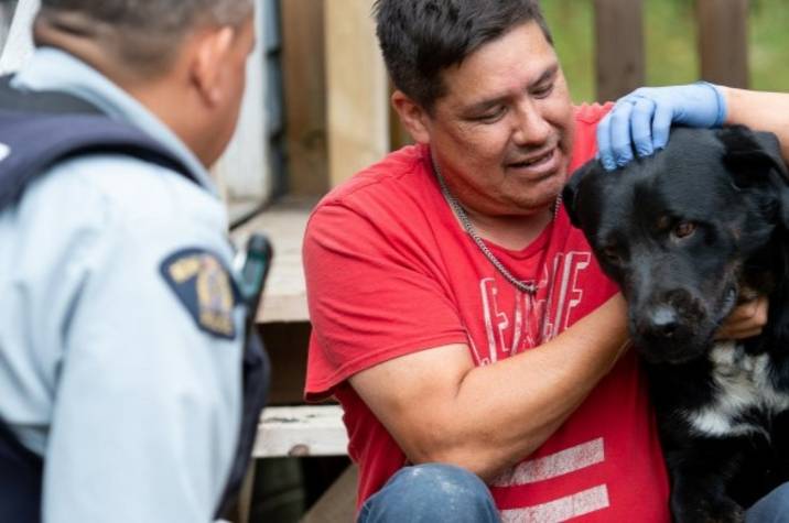 Police officer speaks with seated man embracing a big black dog.